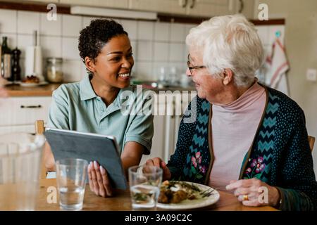 Happy female caretaker holding tablet PC while talking with senior woman having breakfast in kitchen Stock Photo