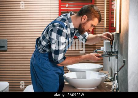 Uses a screwdriver for repairs. Plumber in blue uniform is at work in the bathroom. Stock Photo