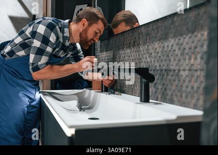 Side view. Fixing water tap. Plumber in blue uniform is at work in the bathroom. Stock Photo