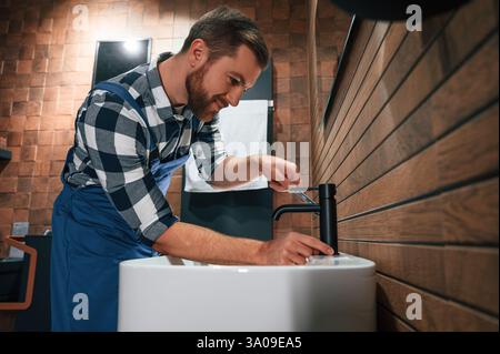 Standing by sink and fixing water tap. Plumber in blue uniform is at work in the bathroom. Stock Photo