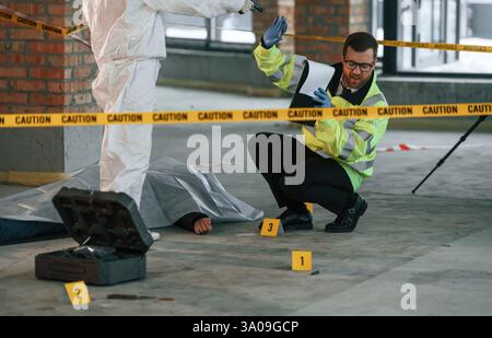 Police worker is sitting on the ground near the dead body while woman is standing with a pistol and threatens him. Stock Photo