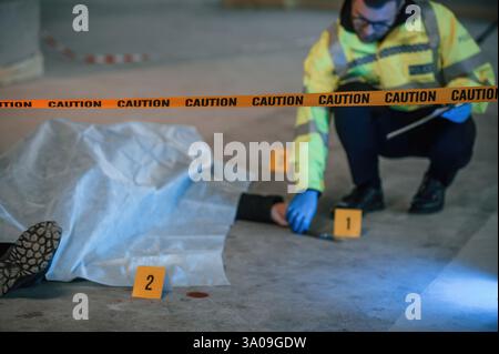 Knife is on the floor. Male detective is collecting evidence in a crime scene near dead body on the construction site. Stock Photo