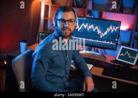Sitting and looking at the camera. Stock trader is working by pc indoors. Conception of exchange and money. Stock Photo