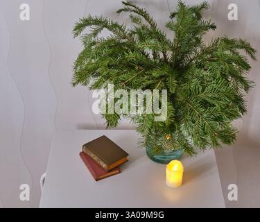 Cozy scene with lush green fir branches in glass vase, two vintage books, and lit LED candle on white table against neutral background. Warm and rusti Stock Photo