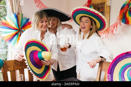 Three cheerful middle-aged Caucasian women wearing colorful Mexican sombreros, laughing and enjoying a festive celebration. Stock Photo