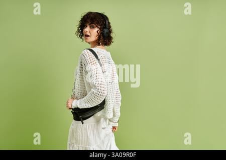 Charming young lady in a stylish outfit enjoys music while standing near a calm green wall Stock Photo