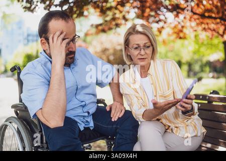 Man in wheelchair is spending time with his mother in park. They are using smartphone and talking. Stock Photo