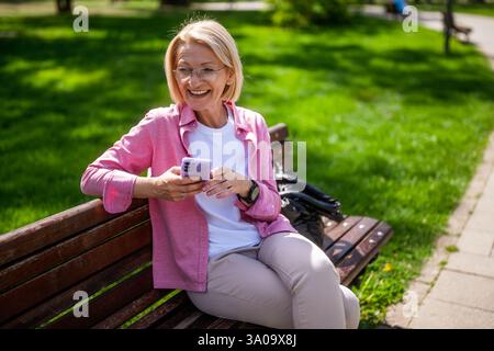 Portrait of happy mature woman in park. She is sitting on bench and using smartphone. Stock Photo