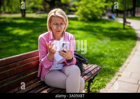 Portrait of worried mature woman in park. She is sitting on bench and having video call on smartphone. Stock Photo
