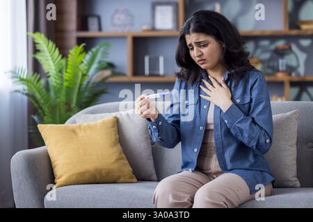 Anxious young woman sitting on a sofa at home, looks worried after taking a pregnancy test uncertain about the future. Stock Photo