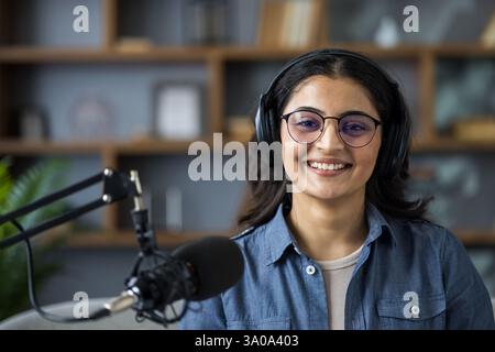 Portrait of a cheerful young woman wearing headphones in front of a microphone, ready to record a podcast or broadcast live from home. Stock Photo