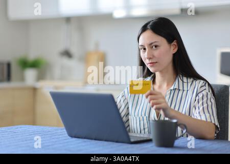 Serious asian woman paying online with credit card and laptop looking at you sitting in the kitchen at home Stock Photo