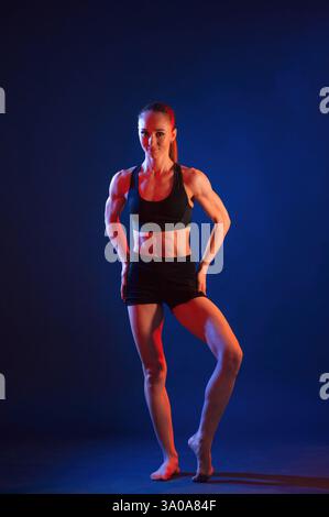 Standing, posing. Beautiful muscular woman is indoors in the studio with neon lighting. Stock Photo