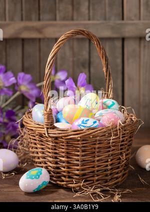 Decorated Easter eggs in a basket with purple daisies on rustic wooden background Stock Photo