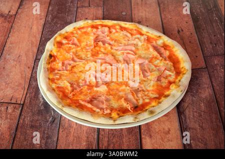 A pizza with ham and cheese on top of a wooden table. The pizza is placed on a white plate Stock Photo
