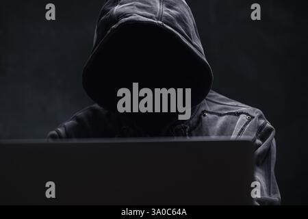 Hooded computer hacker using laptop computer, selective focus Stock Photo