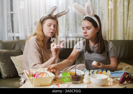 A mother and daughter wearing bunny ears sit on a couch, selecting decorations for Easter eggs. The girl reaches for an ornament from a basket while h Stock Photo