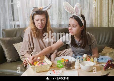 A mother and daughter wearing bunny ears sit on a couch, selecting decorations for Easter eggs. The girl reaches for an ornament from a basket while h Stock Photo