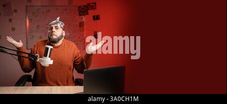 Bearded man in aluminum foil hat can't help, makes gesture of I dont know while Live streaming, presenter with microphone looking at camera Stock Photo