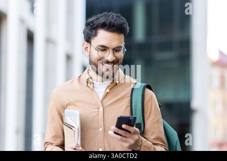 Smiling student with backpack and books checks his mobile phone while on campus, showcasing connectivity and modern education Stock Photo