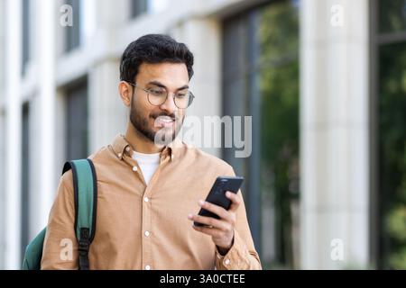 Joyful and happy student walking with phone in hands and backpack, man typing text message and browsing social media. Stock Photo
