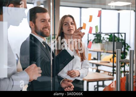 Plans for the project. Group of coworkers in the office is standing together and putting stickers on the glass. Stock Photo