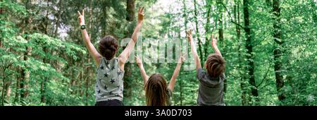 Three children stand in a lush green forest with their arms raised, making peace signs. They are seen from behind, enjoying the outdoors and the fresh Stock Photo
