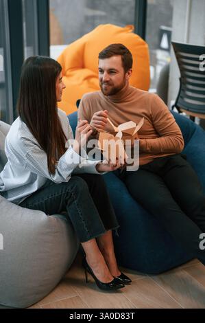 Sitting and talking. Office workers are in the rest zone is having a break and eating food from paper eco boxes. Stock Photo