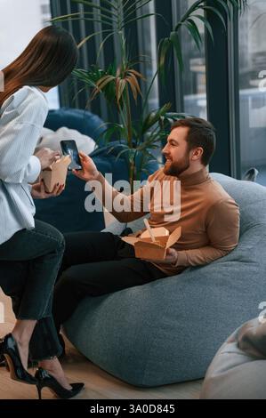 Sitting and talking. Office workers are in the rest zone is having a break and eating food from paper eco boxes. Stock Photo