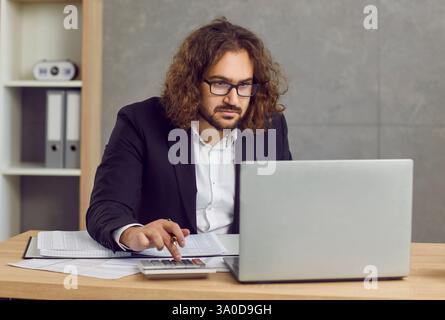 Young focused serious business man working on laptop at office making calculations. Stock Photo