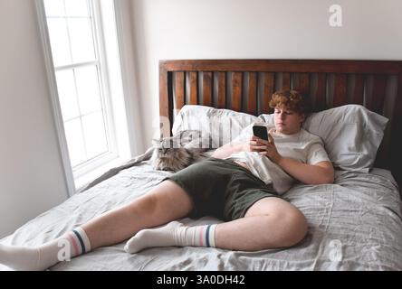 Young man laying on bed with cat using mobile phone Stock Photo