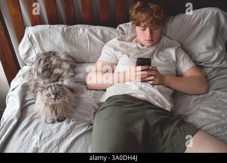 Young man laying on bed with a cat using mobile phone Stock Photo