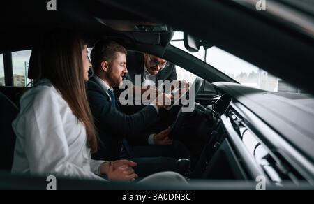 Side view. Car showroom worker is consulting man and woman that are sitting inside of automobile. Stock Photo