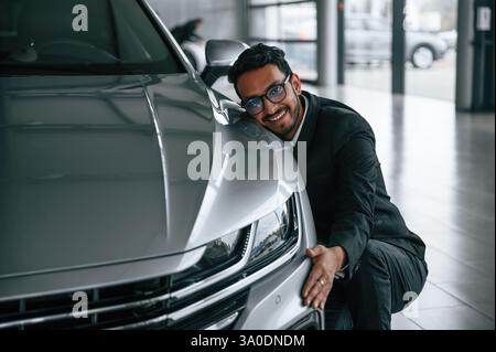 Embracing the transport. Handsome indian man in suit is in the car showroom with the vehicle indoors. Stock Photo