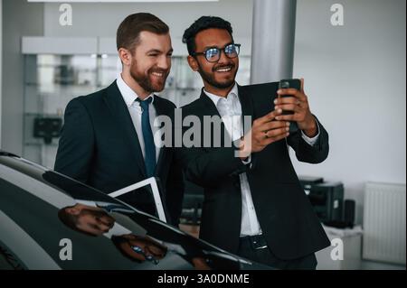Having fun, using smartphone. Two businessmen are working together in the car showroom. Stock Photo