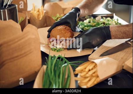 Close up view of burger, French fries and vegetables. Man is packing food into the paper eco boxes. Indoors, restaurant. Stock Photo