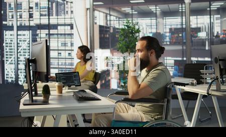 IT worker with disability sitting in wheelchair in office working at computer desk, focused on testing programs. Software engineer with paraplegia in inclusive workplace checking code on PC, camera B Stock Photo