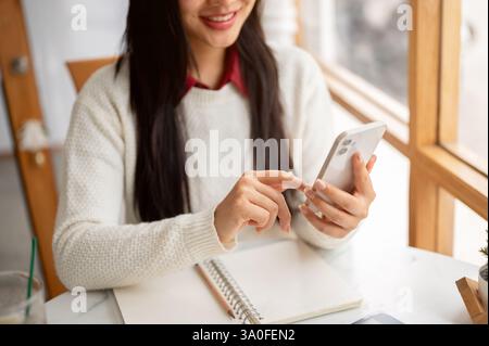 Close up of a smiling asian woman holding and typing on the phone, sitting at the cafe table with notebook and pencil. Chatting with a friend or scrol Stock Photo