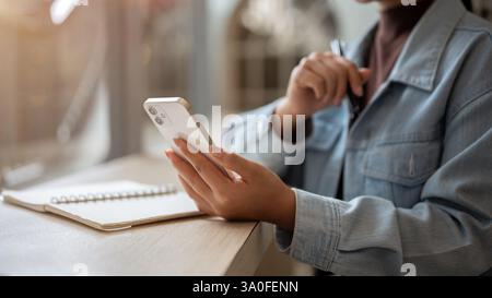 A close up of a person looking at the smartphone holding in left hand with right hand holding a pen while sitting at the wooden table. Watching videos Stock Photo