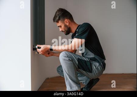 Plumber worker installing heating radiator in empty room of a newly built house. Stock Photo