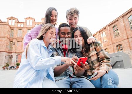 Cheerful group of students browsing with a smartphone, sharing laughter and excitement as they explore a mobile application, showcasing the joy of friendship and connection in the university campus. High quality photo Stock Photo