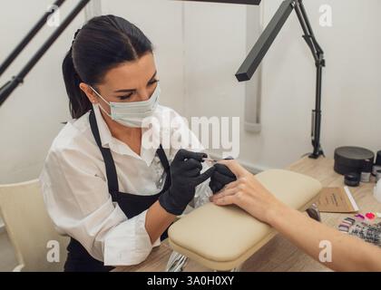 A manicurist in gloves and a protective mask carefully applies varnish to the client's nails in a beauty salon. Professional nail painting procedure Stock Photo