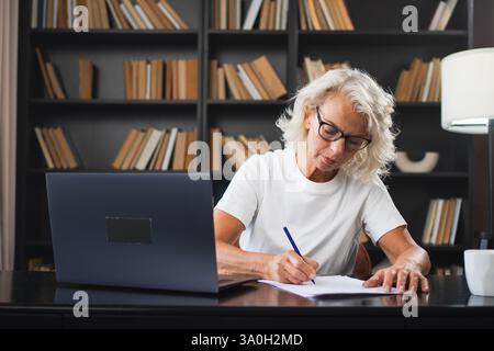 Middle aged senior woman using laptop computer writing notes at workplace. Focused mature old businesswoman work at office making written records doing online work. Mature boss professional worker Stock Photo