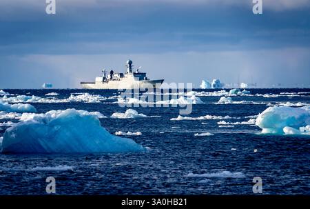 A research vessel navigates through icy waters, surrounded by floating icebergs. The ship is equipped for Arctic exploration, with a cloudy sky in the Stock Photo