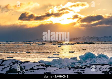 A stunning sunset over icy waters, with floating icebergs and a dramatic sky filled with clouds. The sun casts a golden glow on the landscape, creatin Stock Photo
