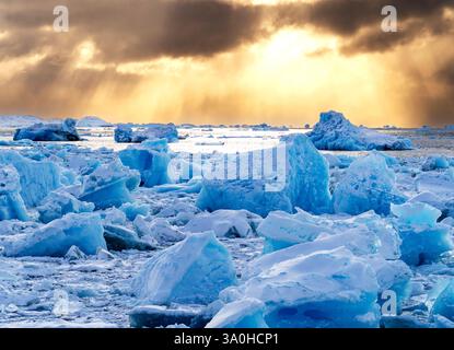 A stunning view of icebergs in a frozen landscape, illuminated by golden sunlight breaking through dark clouds. The icy terrain reflects shades of blu Stock Photo