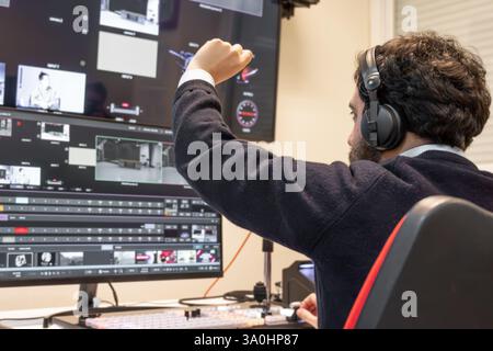 A television director in a broadcast control room, actively guiding a live production. Surrounded by multiple screens, the director wears a headset and gestures towards a video switcher. Stock Photo