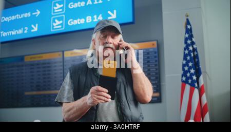 Crowded Airport Terminal: Elderly Man Holding Plane Ticket, Talking by Mobile Phone to Check Flight Information. In the Background, Digital Arrival and Departure Board, Diverse People Walking. Stock Photo