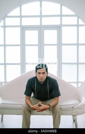 Portrait of an asian man sitting on an elegant couch, indoor with bright window. Looking at the camera. He is stylish and contemplative, conveying con Stock Photo