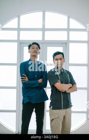 Two Asian men standing confidently indoors near a window with their arms crossed, showcasing teamwork and collaboration. The modern and stylish indivi Stock Photo